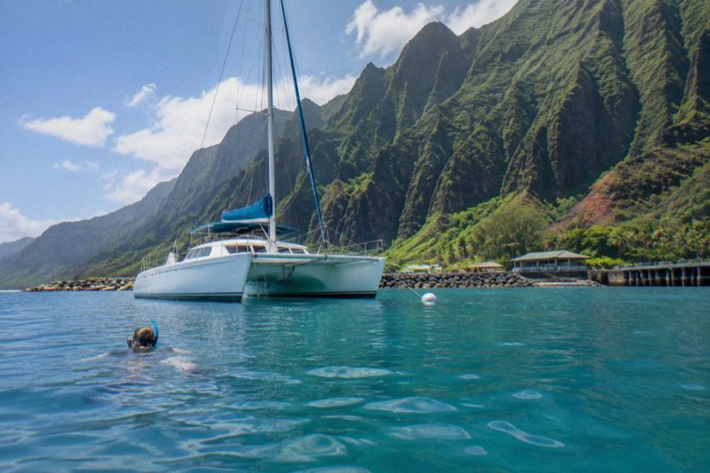 A sailing catamaran anchored near tall emerald cliffs with clear turquoise water below