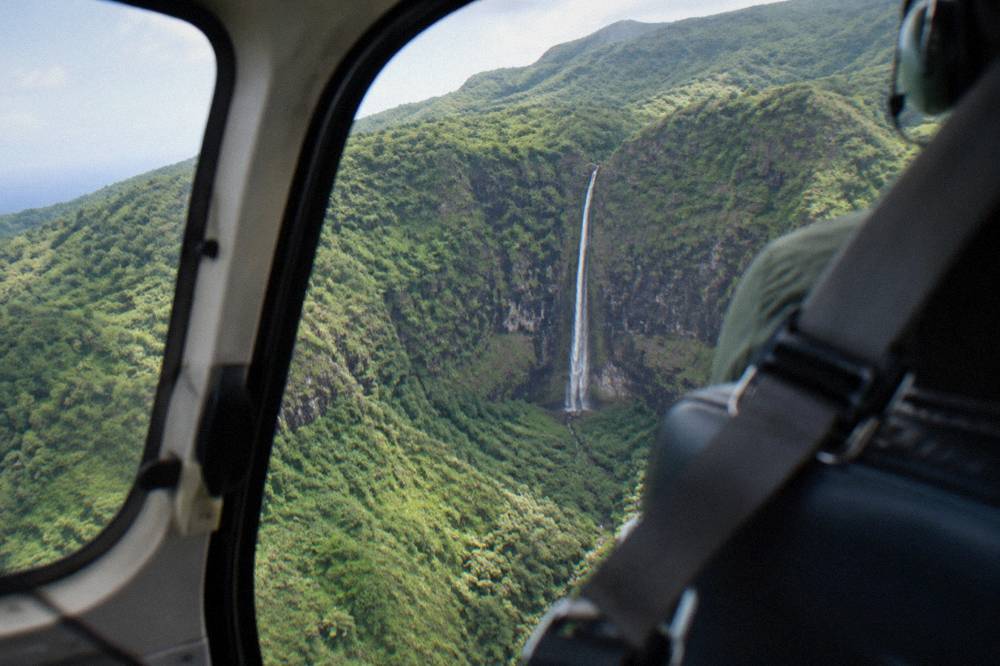 A helicopter cabin view of a long thin waterfall dropping down a dark green cliff face