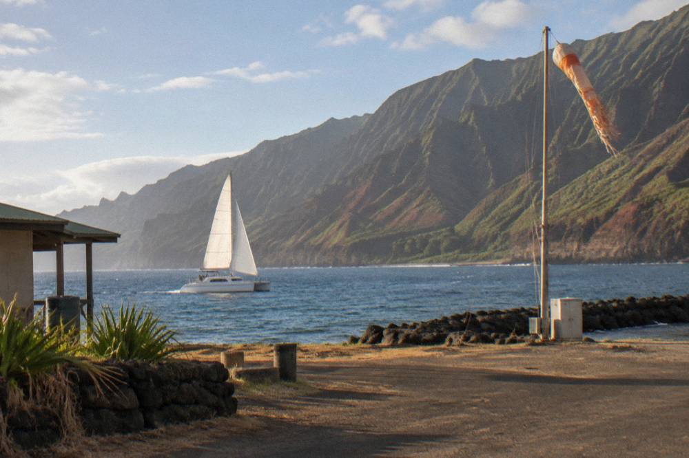 Open catamaran under sail near rugged coastal cliffs in late afternoon