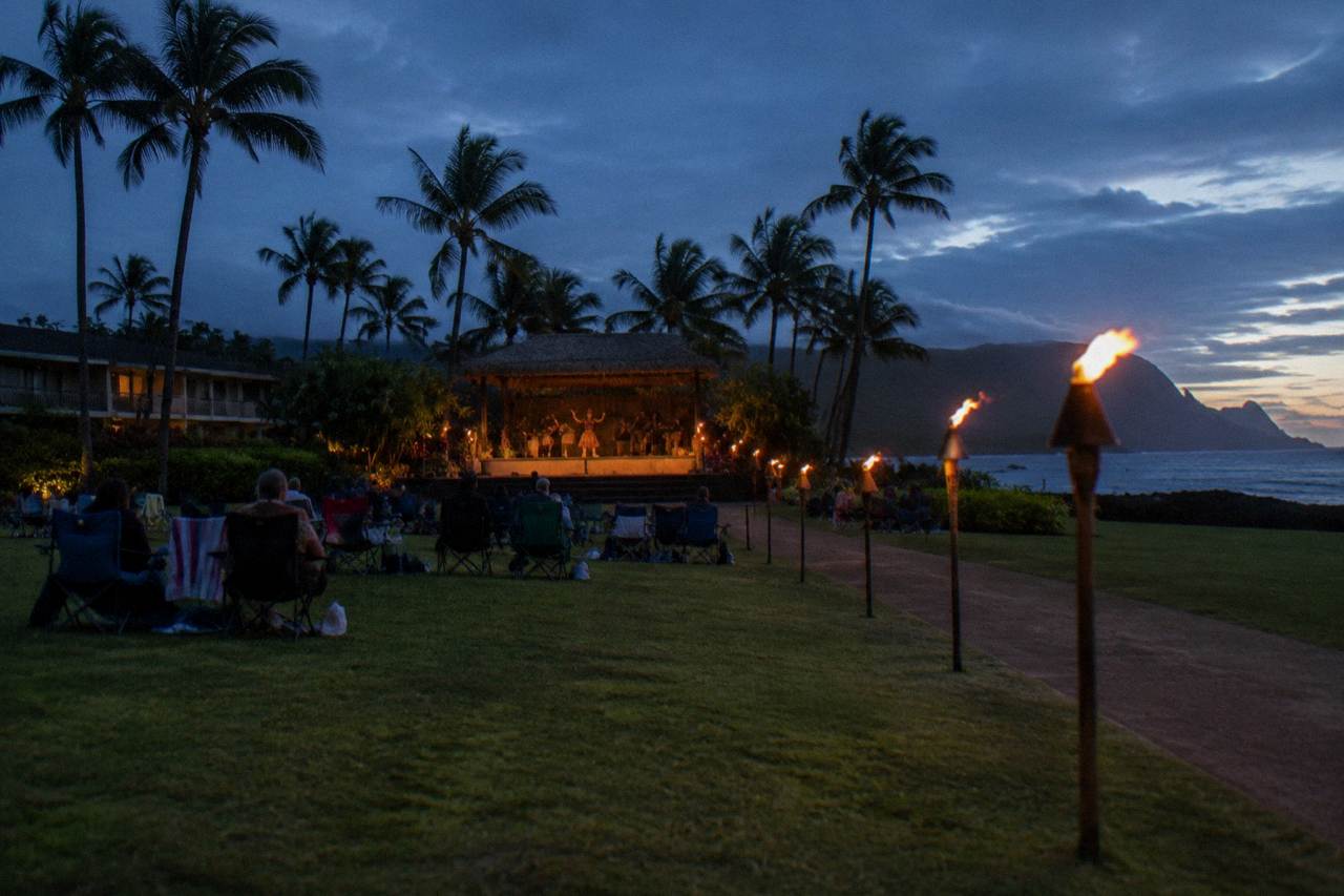 Open-air luau grounds at dusk with tiki torches lit along a lawn toward a palm-lined stage