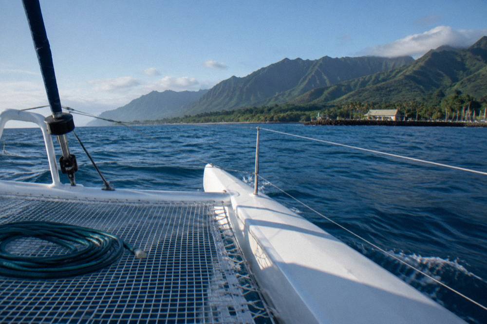 A catamaran under way on open ocean water with a long low island on the horizon and morning light on the swell.