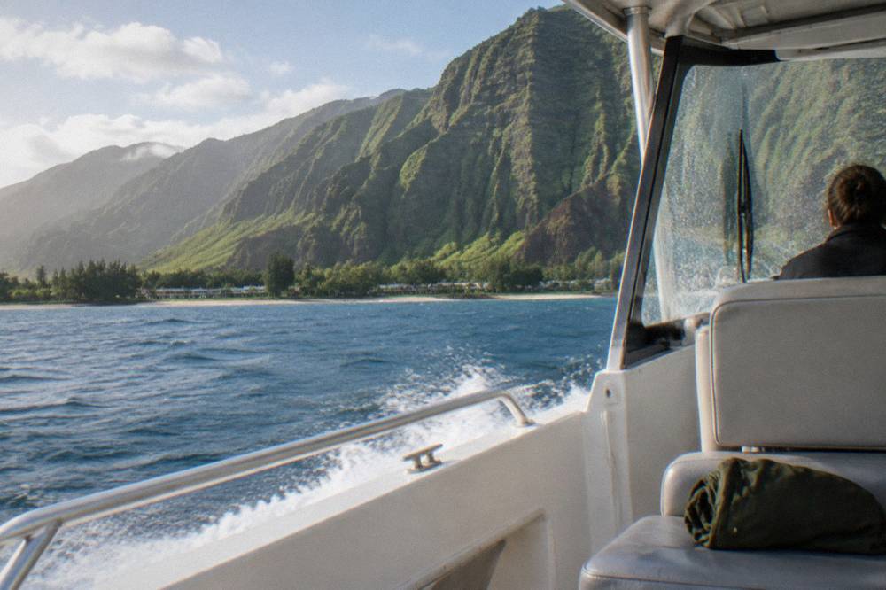 A small power catamaran cruising along a dark green sea cliff coastline in winter light