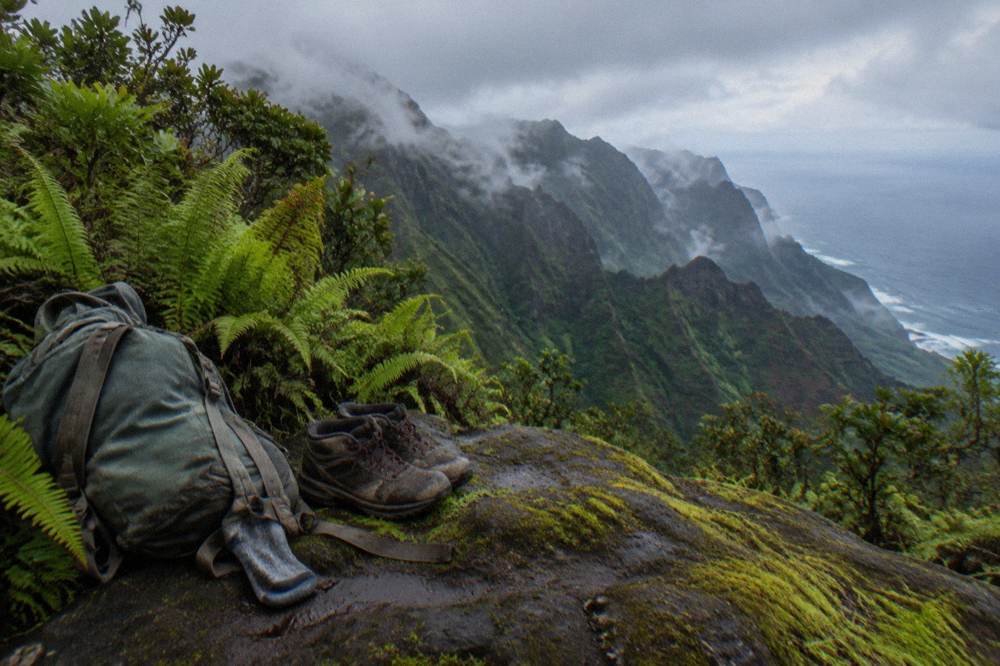 A backpack and hiking boots resting on a rock overlooking a green valley and waterfall