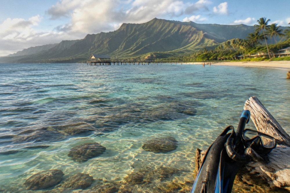 Shallow turquoise lagoon over coral reef with mountain peaks rising behind a sand beach