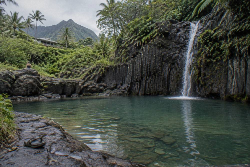 A narrow ribbon of water falling down a dark lava rock face into a clear jungle pool