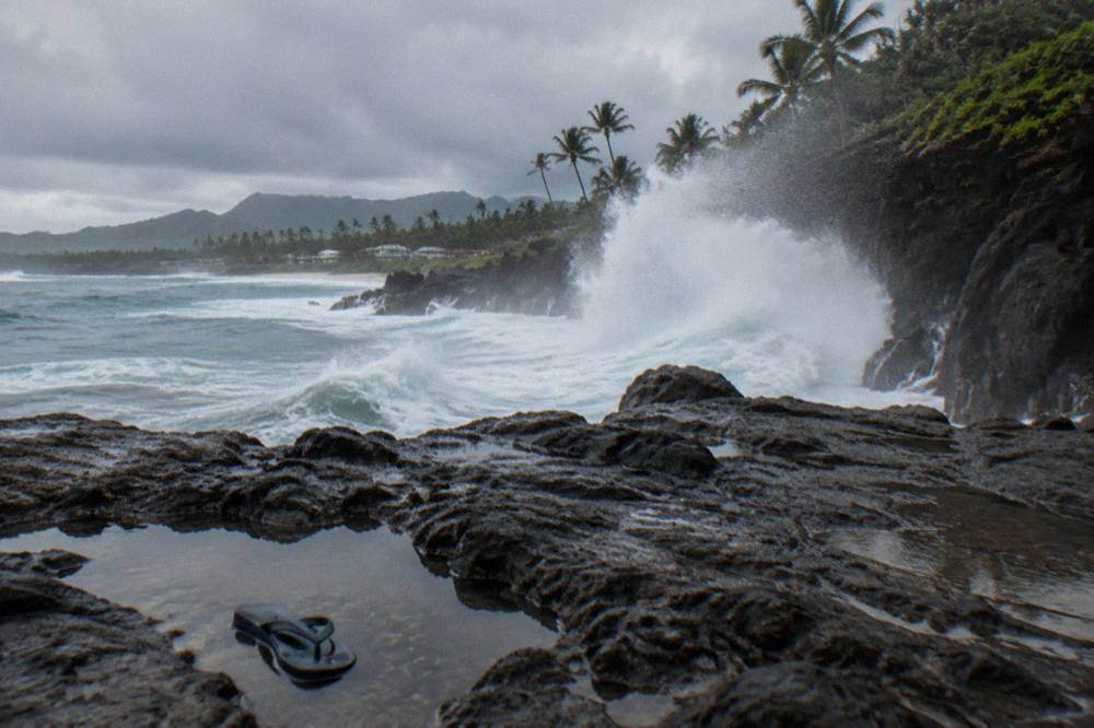 Large ocean waves breaking against a rugged north shore cliff face under a grey sky