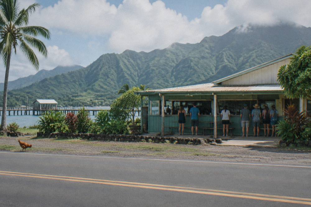A small Hanalei town storefront with a line of people waiting outside a lunch counter under green mountain cliffs