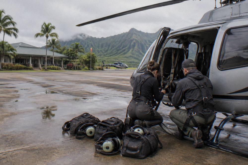 A helicopter safety briefing area with crew checking harnesses beside a parked aircraft