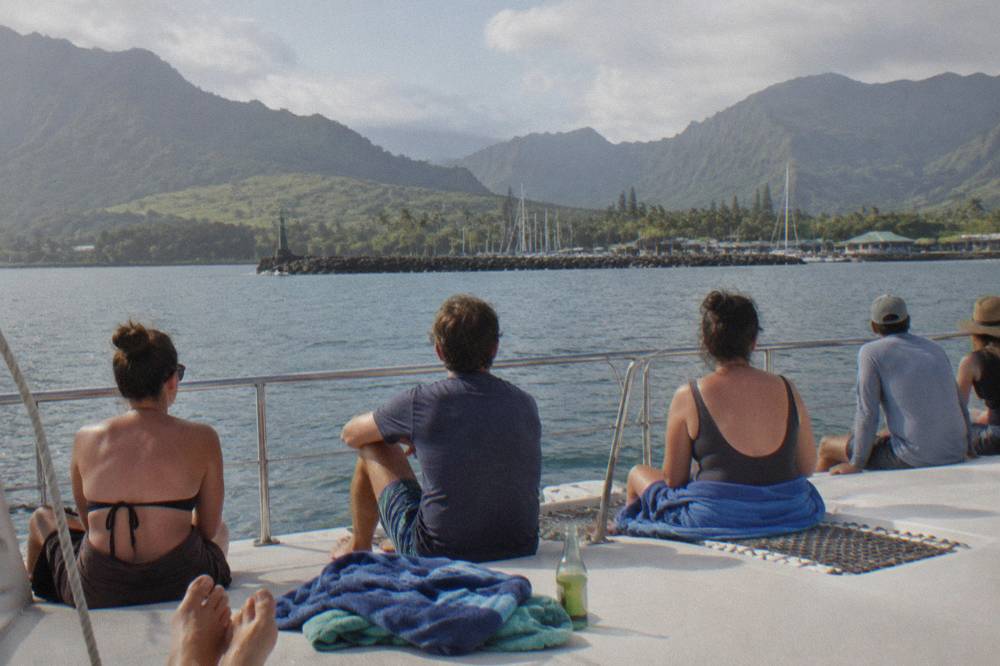 A wide catamaran deck seen from the stern with passengers seated along the rail on calm blue water
