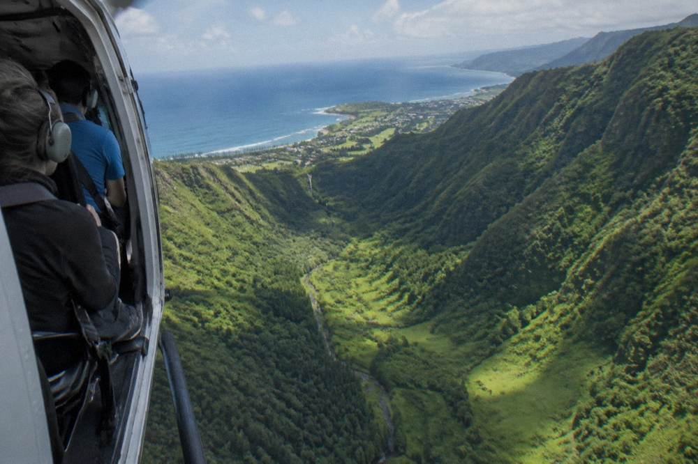 A small helicopter with no doors flying over a narrow green valley