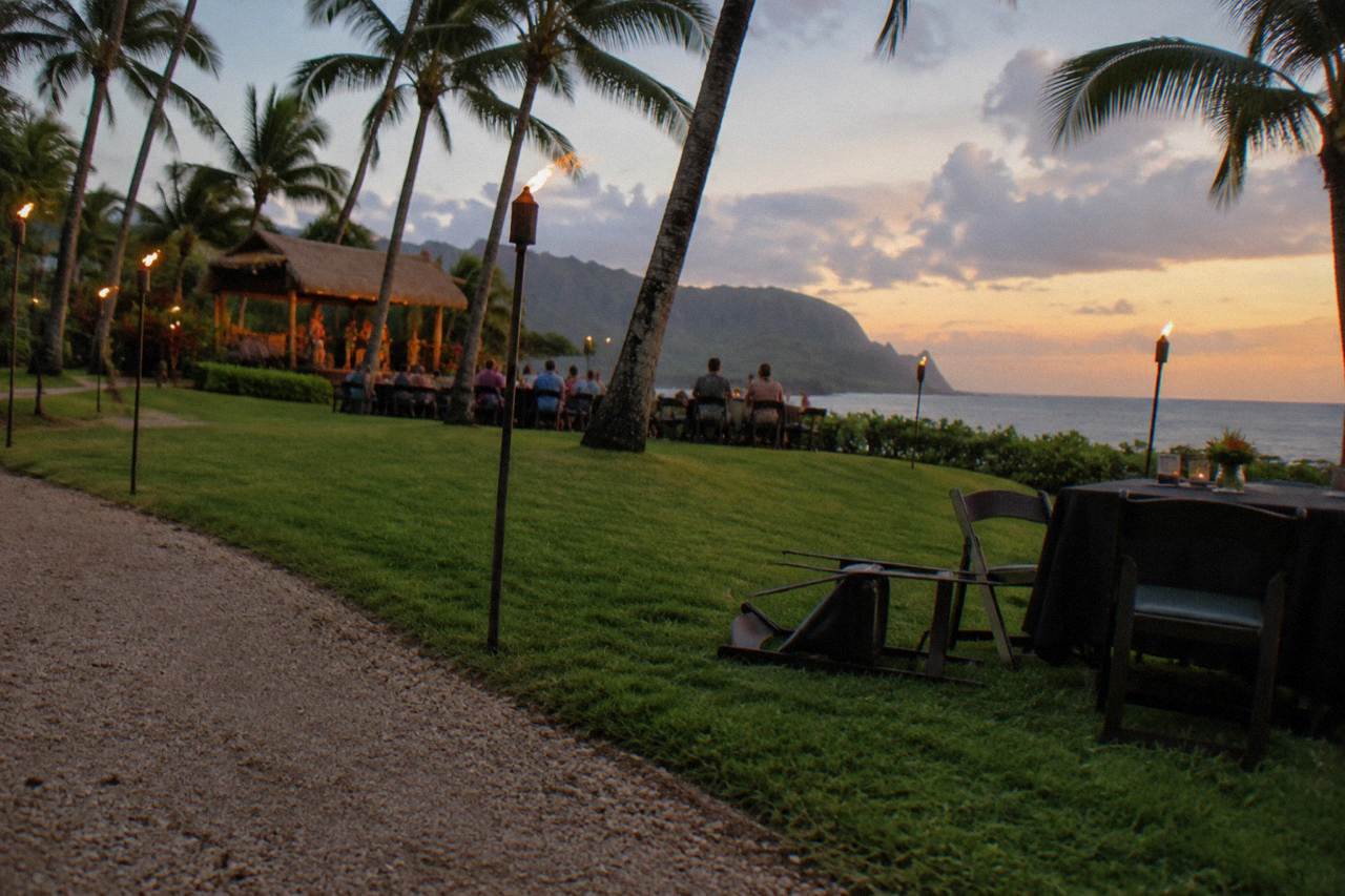 Open-air luau grounds at dusk with tiki torches lit along a lawn toward palm trees