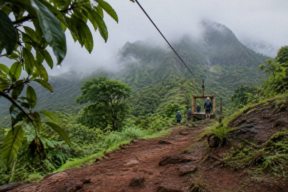 A zipline cable disappearing into misty green mountains under broken clouds