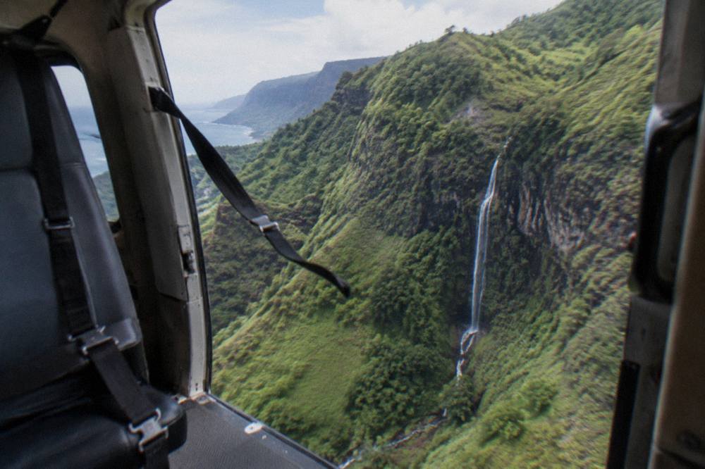 Open door of a helicopter in flight above a green valley with a waterfall