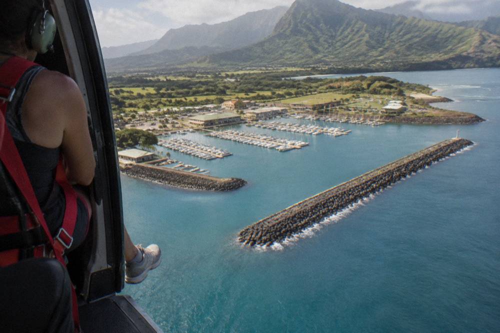 A passenger in a flight harness seated beside an open helicopter door above the ocean