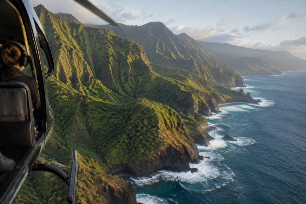 Helicopter flying along a coastline with tall green sea cliffs in morning light