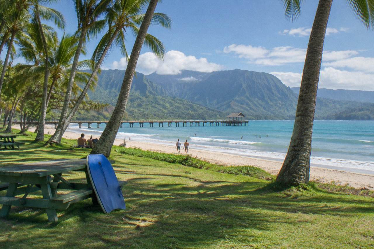 A wide sandy beach with gentle waves, palm trees and a grassy park behind, on a sunny day on Kauai