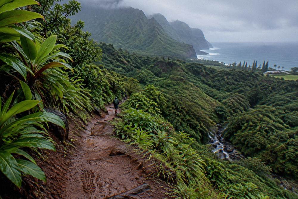 A hiker's view of a red dirt path dropping toward a stream valley