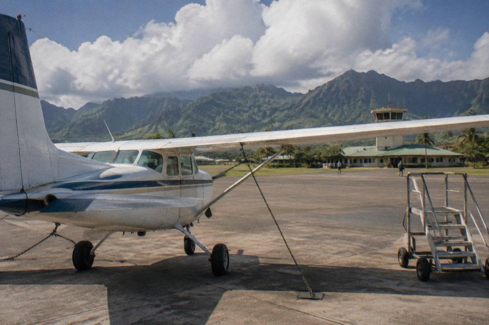 A high-wing sightseeing airplane parked on a runway with green mountains behind it