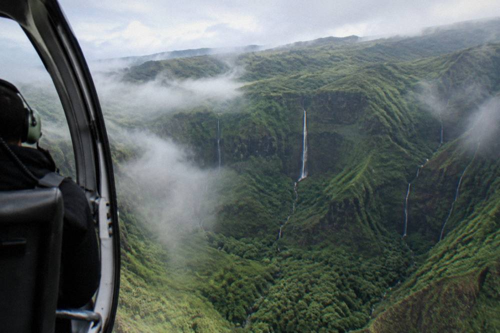 A helicopter banking over a deep green valley with thin waterfalls on the cliff walls