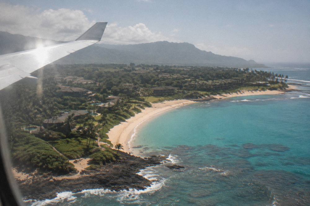 An airplane wing above turquoise water and a curving white sand beach on Kauai