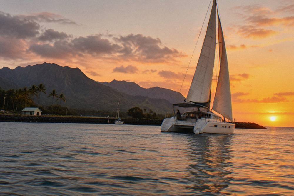 A sailing catamaran heeling slightly with an orange sunset sky above calm water.