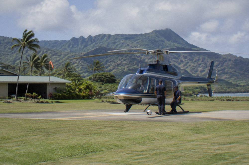 A tour helicopter on a grass apron at a Lihue heliport with rotors stopped and crew standing beside it.
