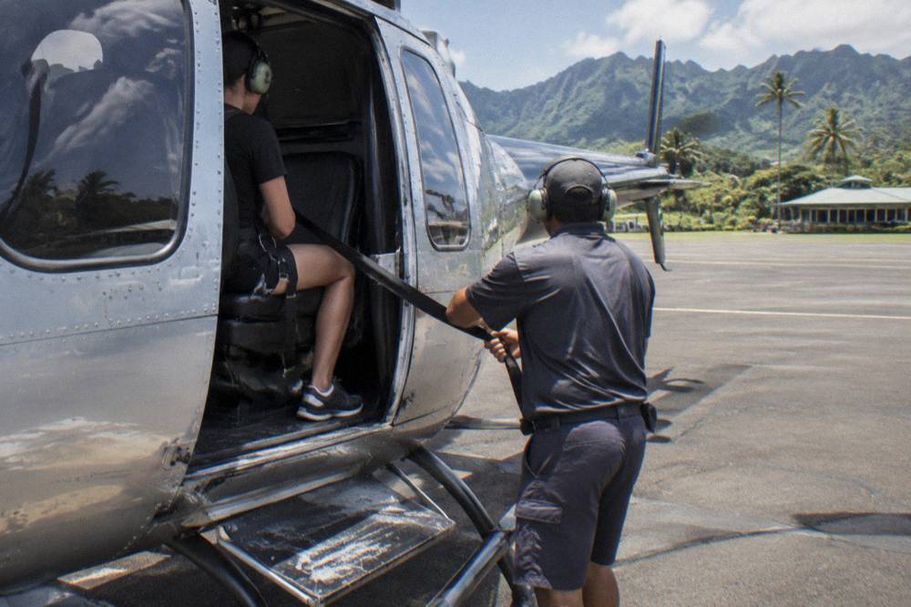 A passenger in a full harness seated in an open helicopter door with a crew member checking straps.