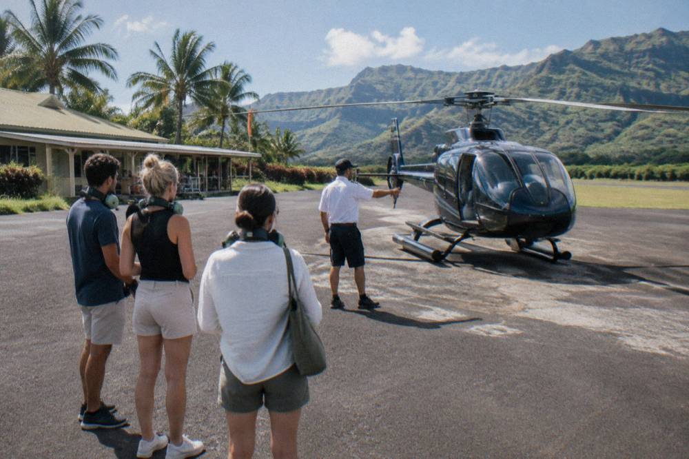 A flight briefing on the tarmac with a pilot pointing at a helicopter while passengers listen wearing headsets around their necks.