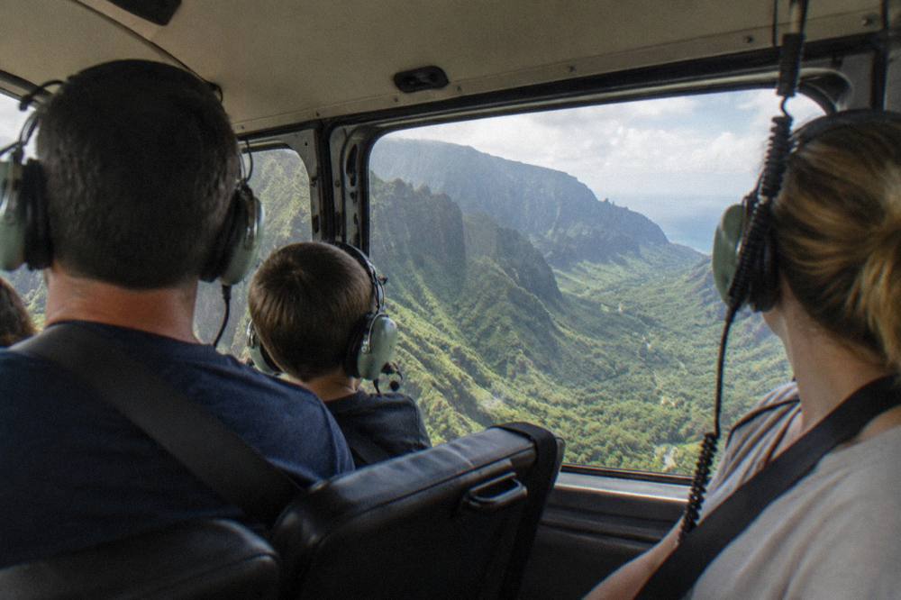 A family in seat belts inside an enclosed helicopter cabin wearing headsets and looking out the windows at green cliffs.
