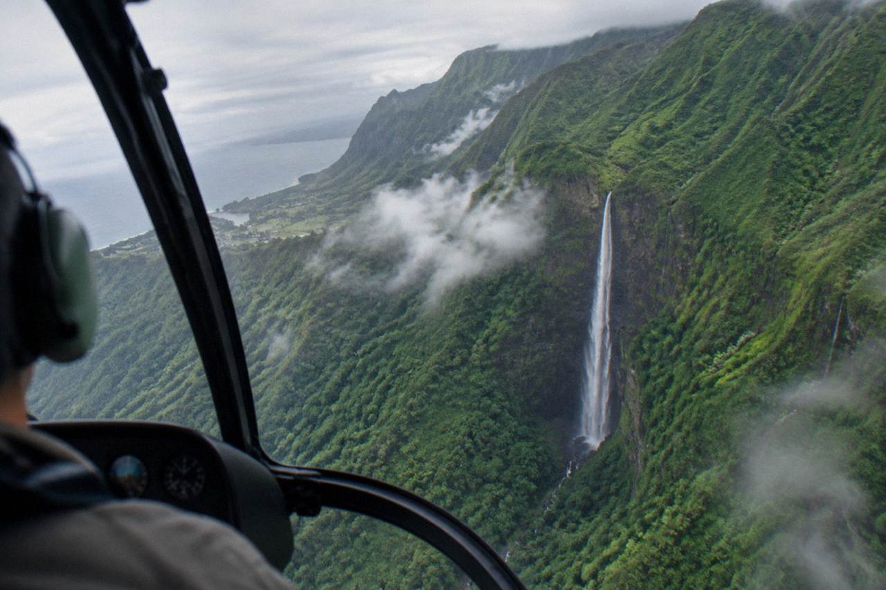 A helicopter flying over green Kauai ridgelines and a tall ribbon waterfall under scattered clouds.