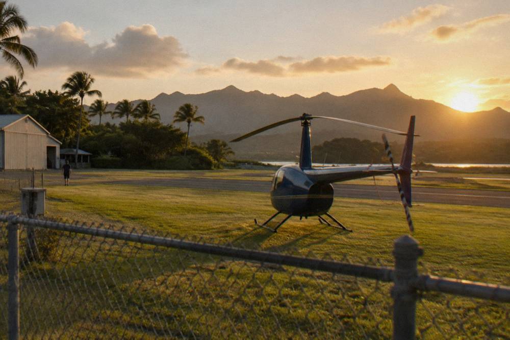 A small helicopter parked on a grassy pad near a runway at golden hour
