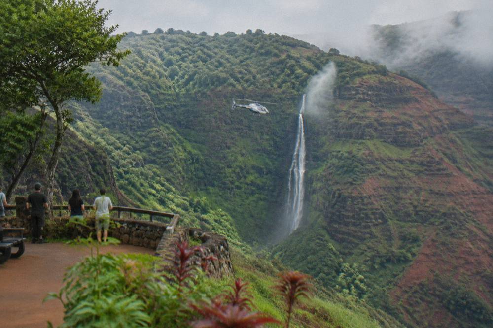 A helicopter hovering beside a tall thin waterfall on a green cliff face