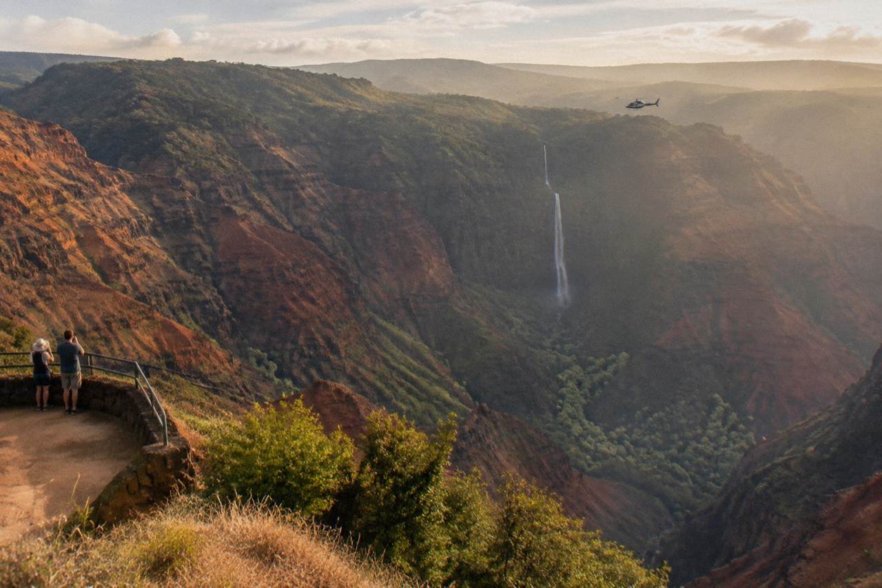 A helicopter flying over green Kauai ridges and a tall ribbon waterfall