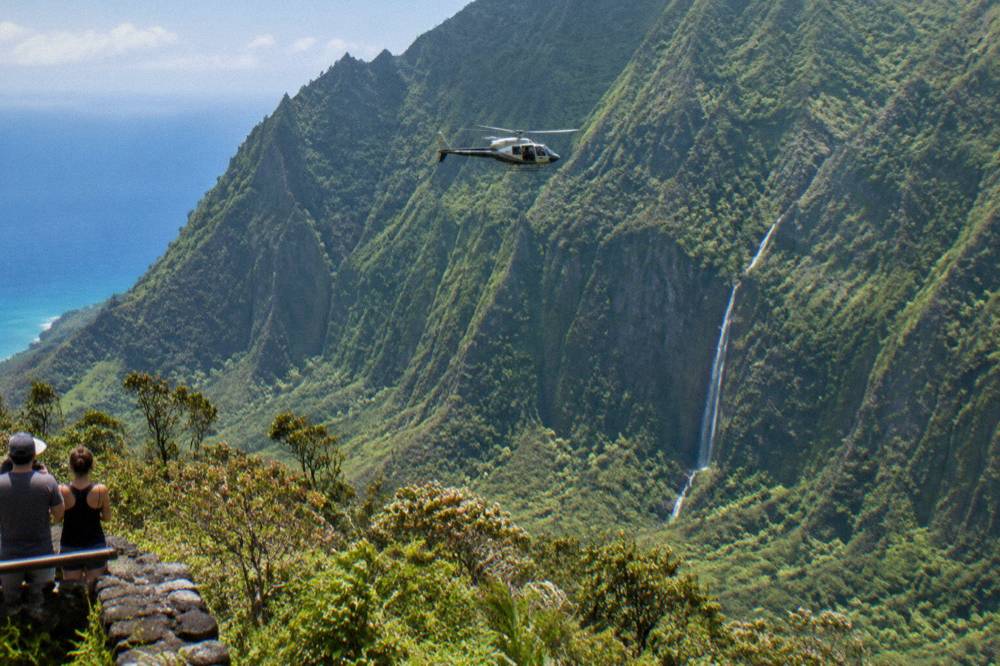 A small helicopter flying doors-off above a deep green canyon with a ribbon waterfall