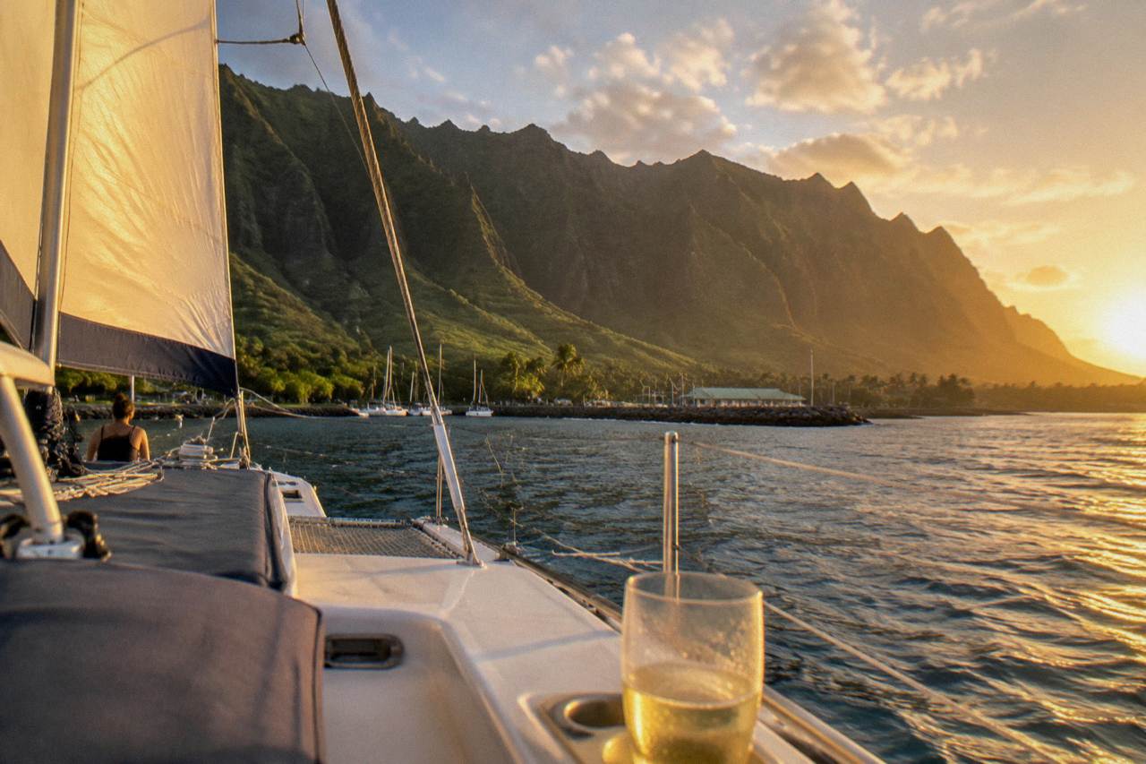 A catamaran under sail along a steep green sea cliff coastline at golden hour