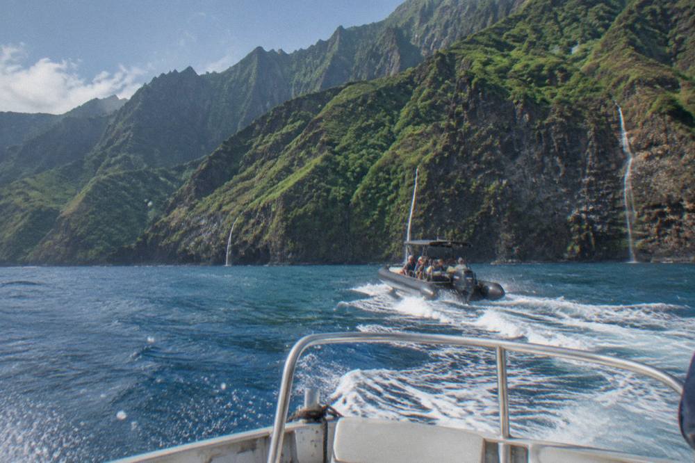 A small boat cruising along tall green sea cliffs with waterfalls trailing to the ocean