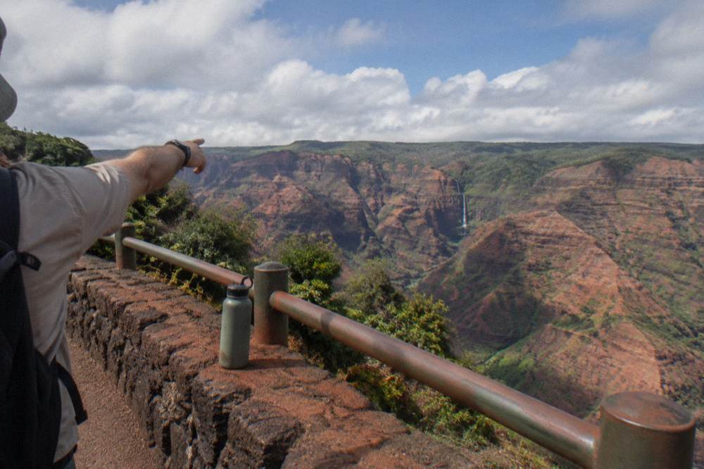Tour guide pointing toward a deep red canyon gorge under scattered clouds