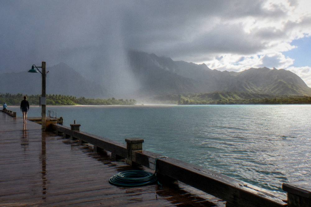 A rain shower passing over the ocean near a green Hawaiian shoreline.