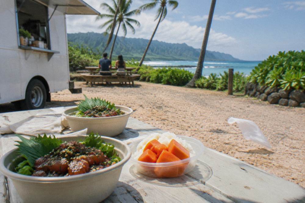 Plates of poke, rice and tropical fruit arranged on an outdoor table beside a food truck