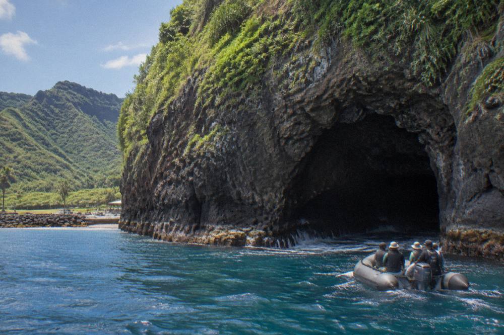 A zodiac raft drifting near a dark sea cave at the base of tall green sea cliffs