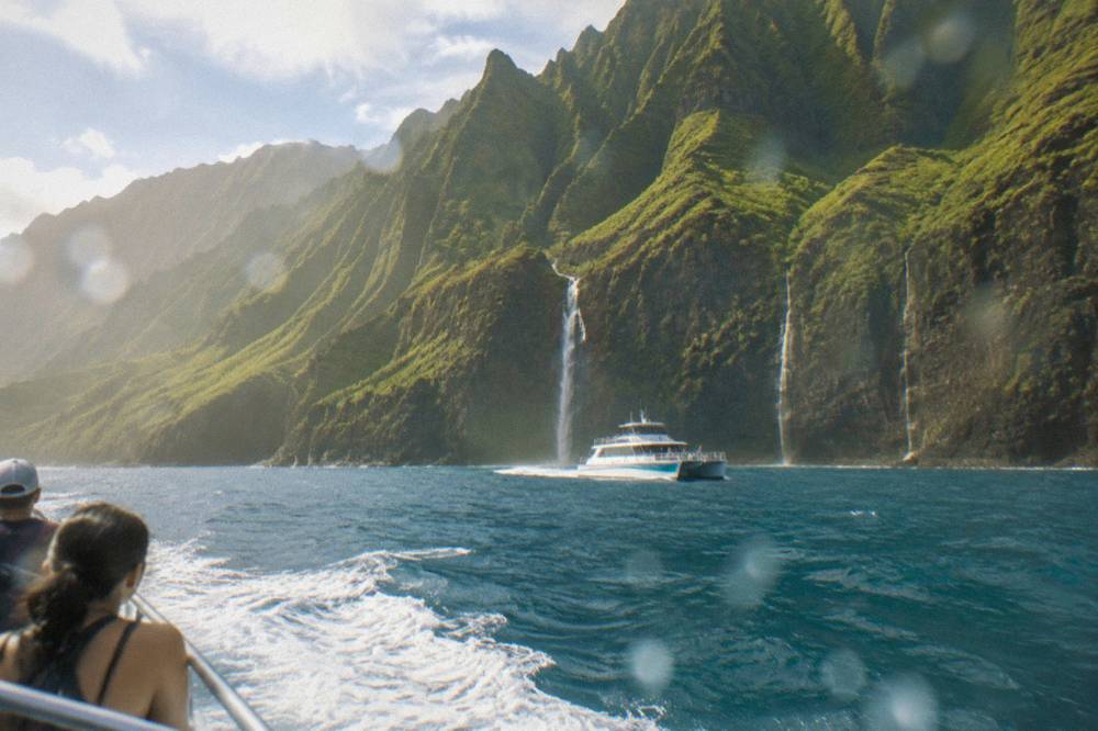 A power catamaran cruising along tall green sea cliffs with waterfalls streaking the rock face