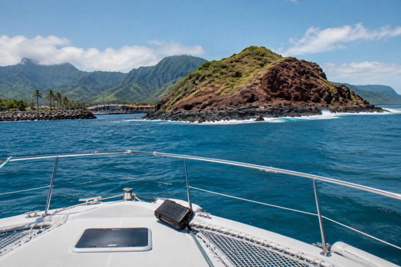 Clear turquoise water washing over a crescent-shaped rocky islet off a distant island coastline under scattered clouds