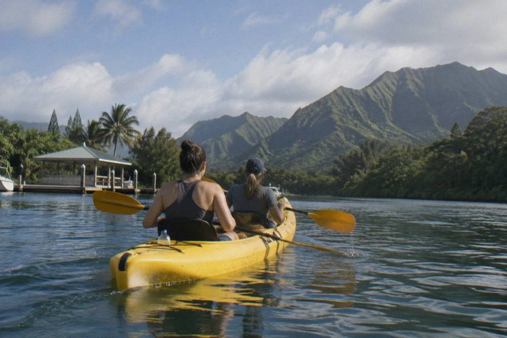 Two people in a tandem kayak paddling on a wide calm river between green forested slopes