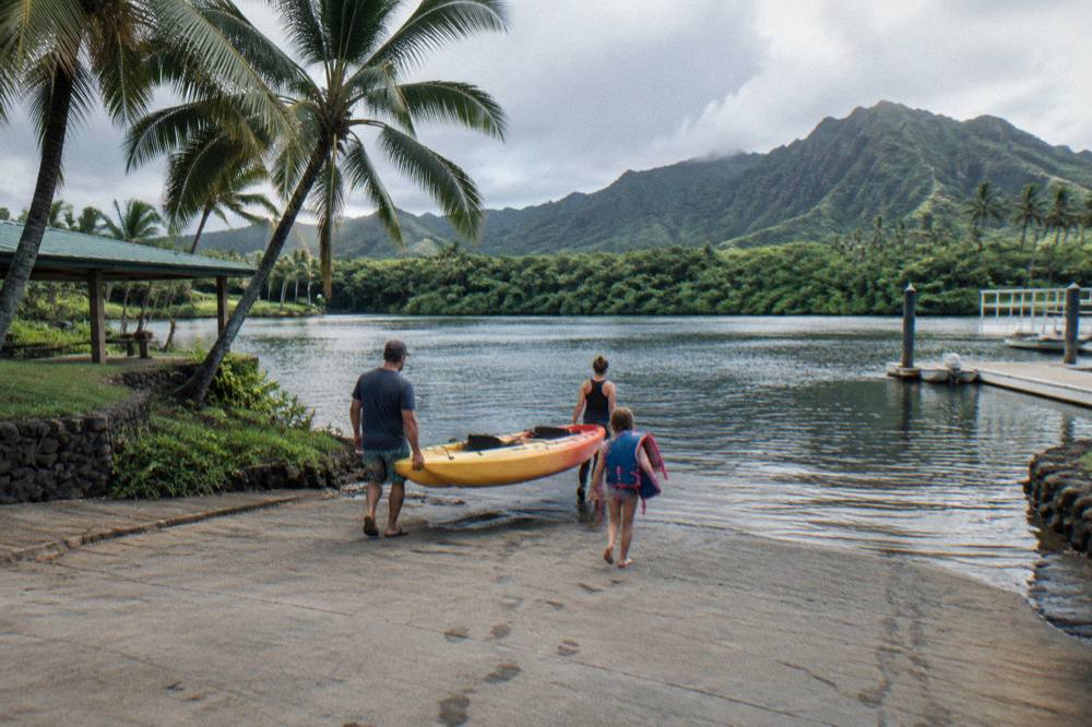A family carrying a tandem kayak toward the water past palm trees at a river landing